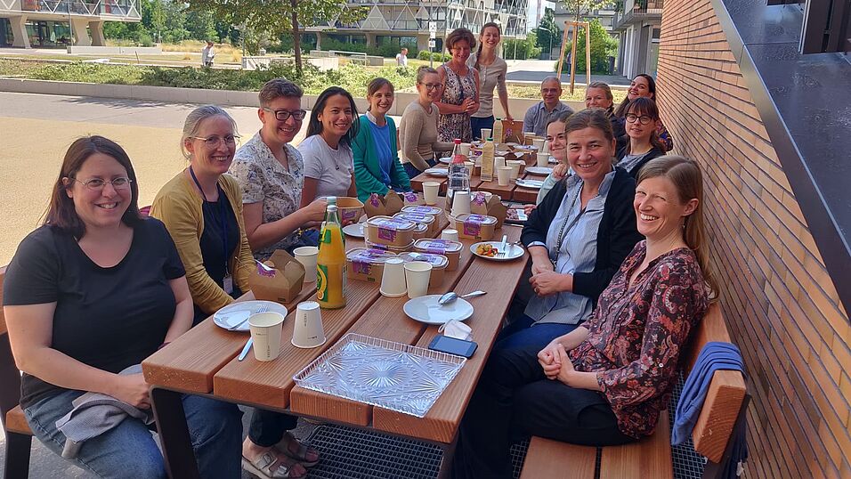 Smiling WoBio members are seated on benches on each side of two long tables (8 left, 6 right). The speaker (Kevin Laland) is seated at the end of the table. The leftovers from a meal are on the table. The bench on the right is close to the brick wall of the university biology building. In the background, two people are walking in a garden. 