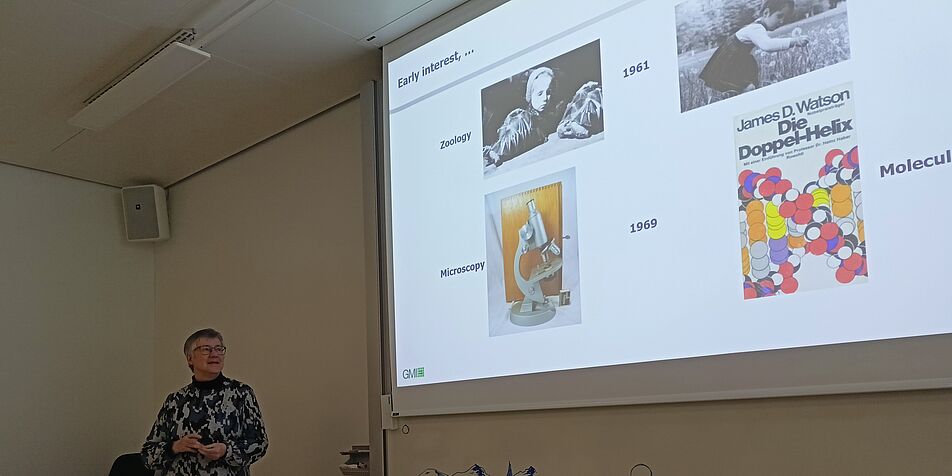 Dr. Ortrun Mittelsten Scheid stands on the lower left corner of the picture. Only the upper part of her body is visible. She is looking at the slide projected on a screen. 
The slide has a white background, and blue writings, with the title: "Early interest". 
There are four pictures on the slide. The two upper pictures are pictures of Ortrun Mittelsten as a little girl, and on the bottom an old-fashioned microscope (left) and a poster of a double helix with the title "James D. Watson - Die Doppel-Helix" (right). 