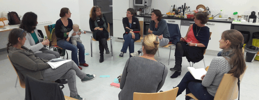 Nine WoBio members are discussing, seated on chairs arranged in a circle. The third woman on the left is holding a baby facing the audience. 